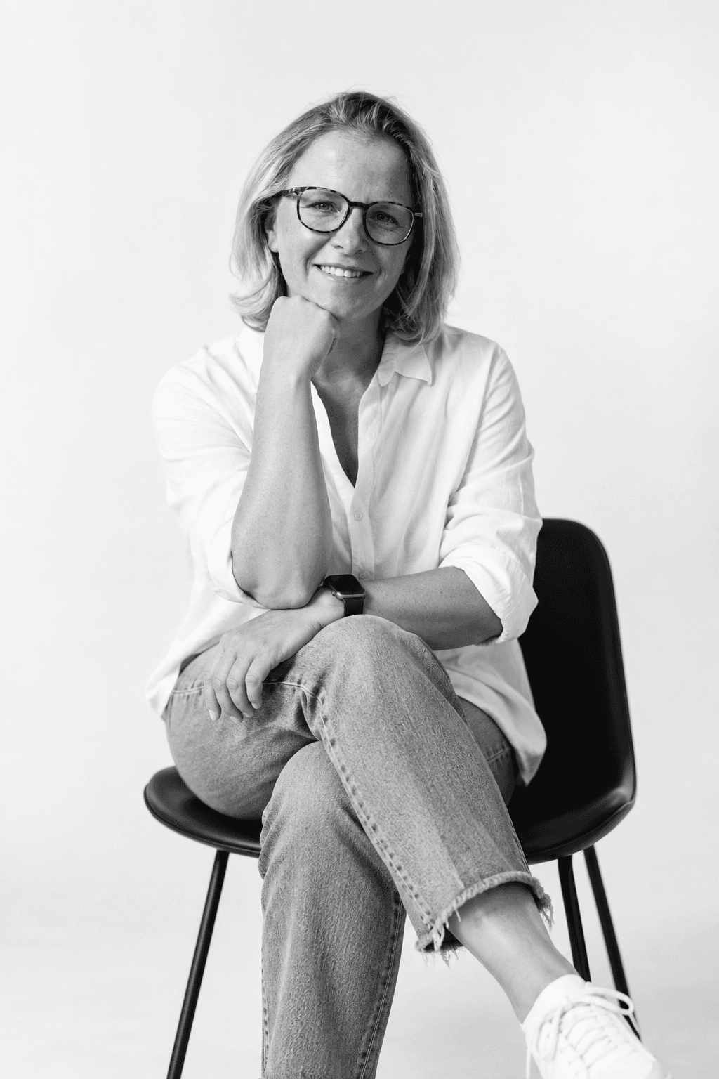 Katka Šumpíková v bílé košili a džínách sedí na židli ve fotografickém ateliéru na světlém pozadí.